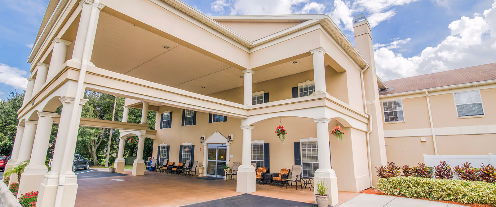 Exterior view of a senior living facility named Madison at Oviedo, showing a covered entrance with columns, outdoor seating with chairs and tables, hanging flower pots, and a clear sky with clouds.