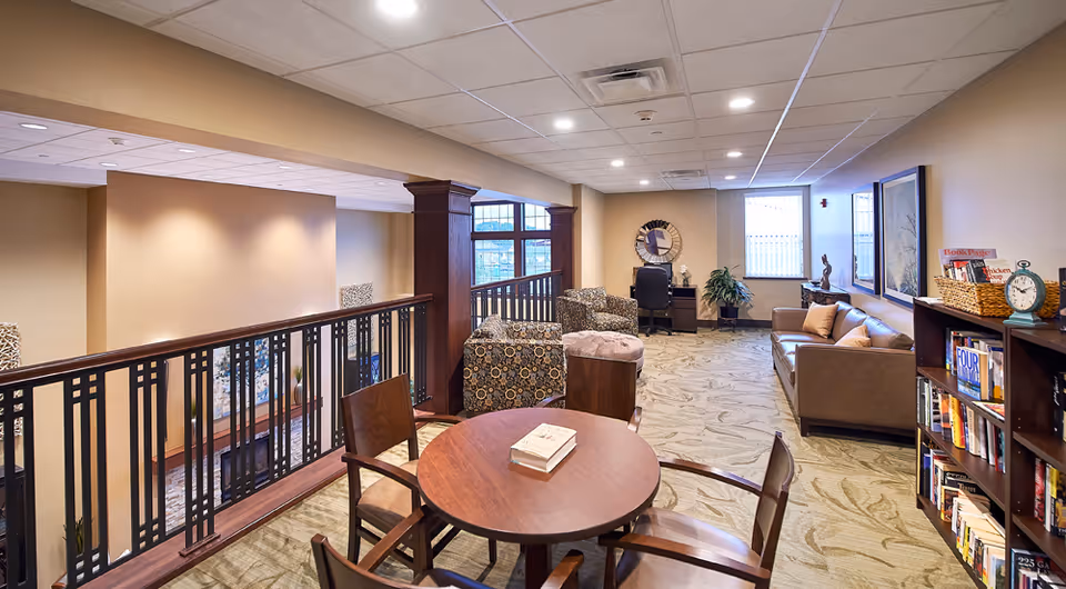 Well-lit senior living lounge with a round table and chairs, armchairs, a sofa, bookshelf, and a desk overlooking an interior atrium.