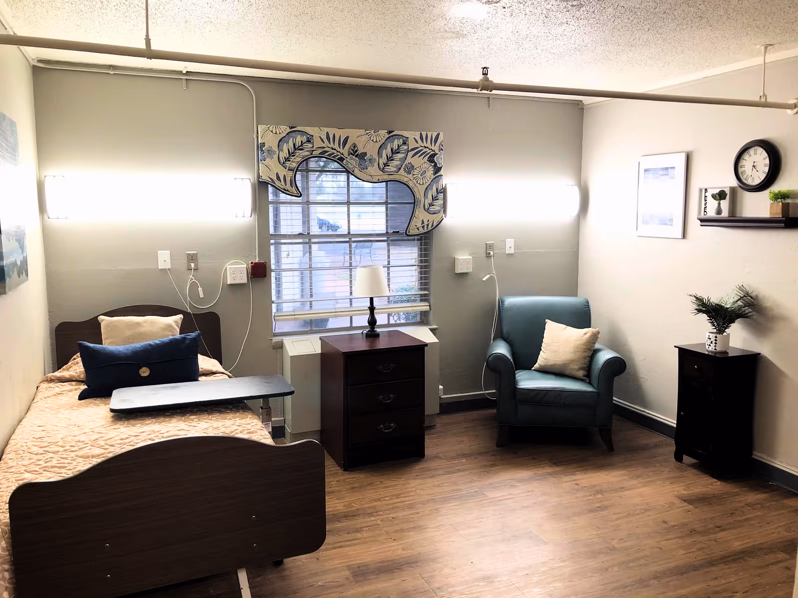 A small, neatly arranged room with a single hospital-style bed on the left side, featuring a beige quilt and two pillows. Next to the bed is a dark wooden nightstand with a lamp on top, positioned in front of a window with blinds and a decorative valance. On the right side of the room is a blue armchair with a beige pillow, a small dark wooden cabinet with a potted plant, and a wall-mounted shelf holding a clock, framed picture, and small decor items. The room has light-colored walls and wood flooring.
