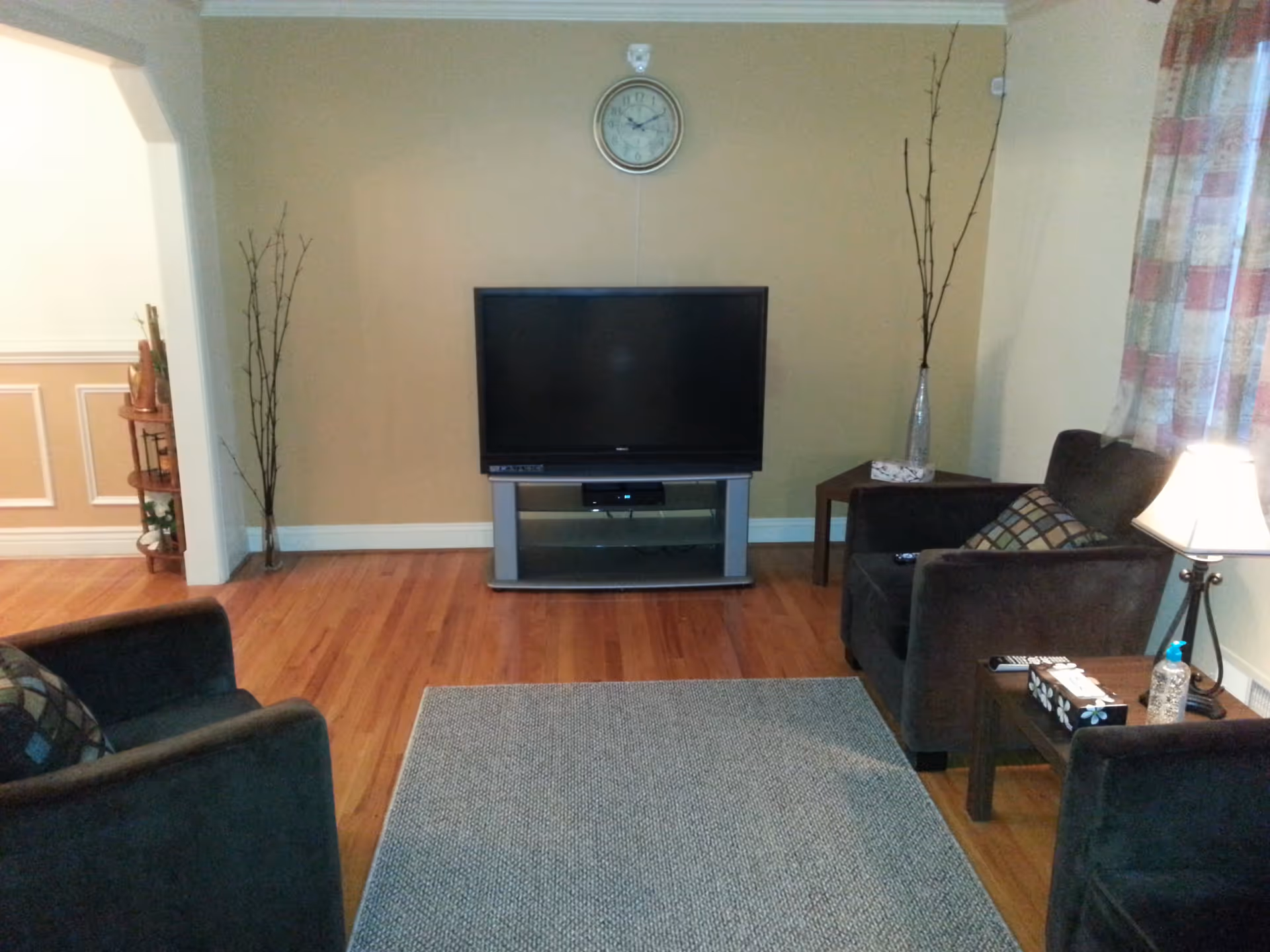 A cozy living room with two dark armchairs facing a flat-screen TV on a stand. There is a clock on the wall above the TV, a small side table with a lamp, a tissue box, and a bottle of hand sanitizer. The room has wooden flooring, a gray area rug, and light-colored walls with a window covered by patterned curtains.