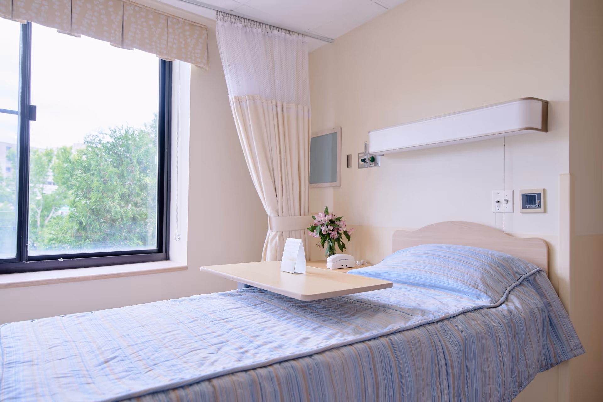 A single bed in a room with light blue striped bedding, a wooden headboard, and a light wooden overbed table. There is a window with beige curtains and a view of green trees outside. A small bouquet of flowers and a white telephone are on a small bedside table next to the bed. The walls are light-colored and there are medical equipment and controls mounted on the wall above the bed.