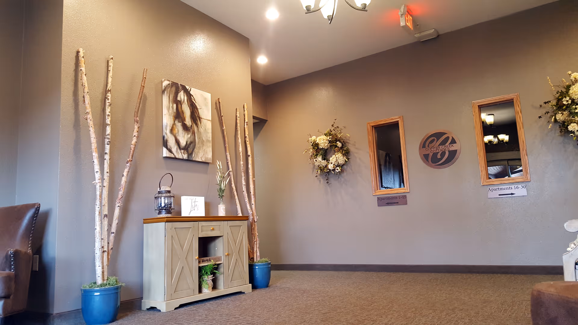 Interior view of a hallway or lobby area with beige walls and carpeted floor. There is a small wooden cabinet with decorative items on top, including a lantern and a small plant. Tall birch branches in blue pots flank the cabinet. On the wall above the cabinet is a painting of a horse. Two rectangular mirrors and a circular Copper Creek logo are mounted on the opposite wall, along with signs indicating apartment numbers. A floral wreath is also hanging on the wall.
