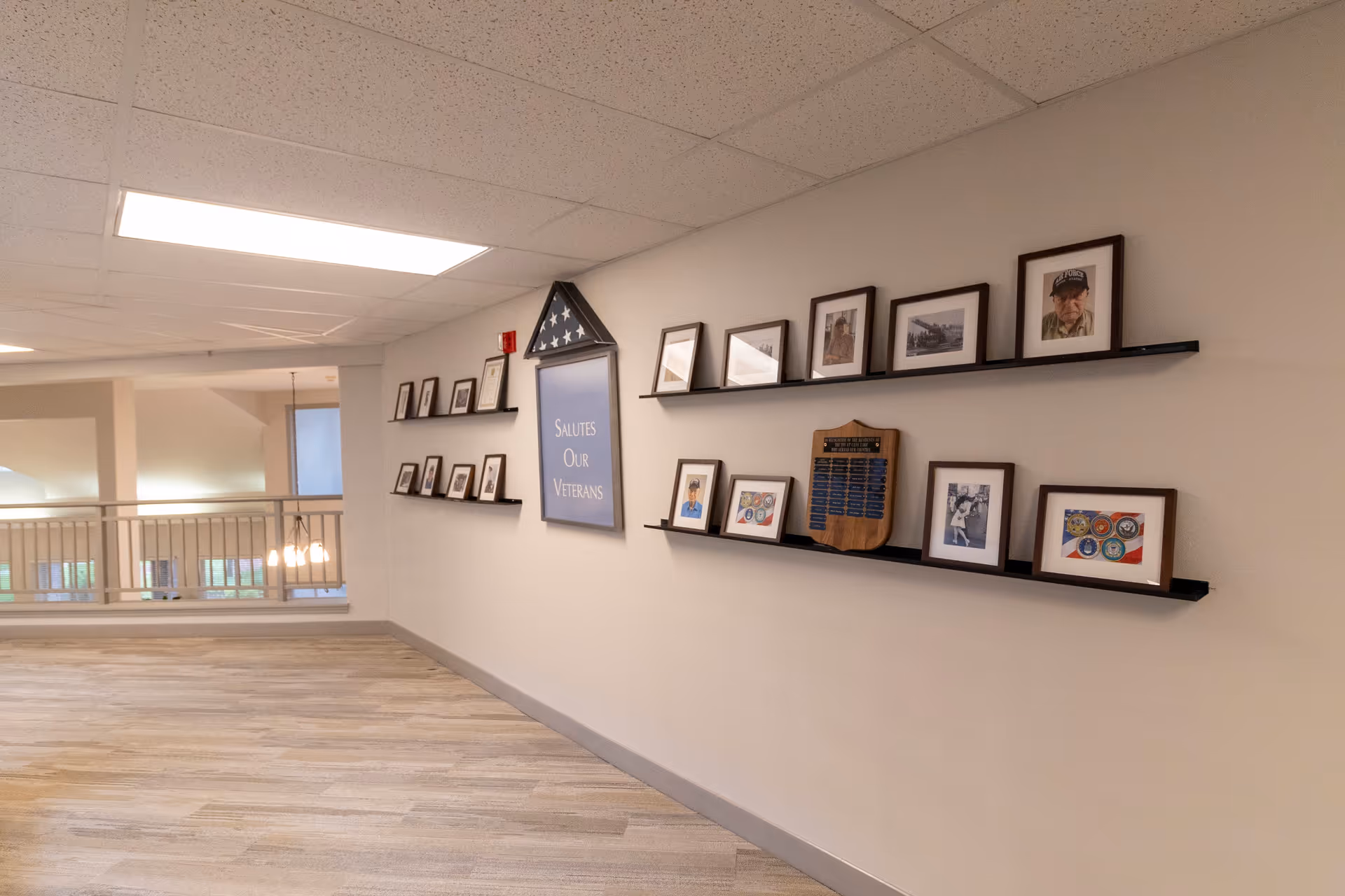 Interior hallway with light wood flooring and white walls featuring a display of framed photographs and a folded American flag in a triangular case. A sign reads 'Salutes Our Veterans'. The hallway has a railing on the left side and ceiling lights.