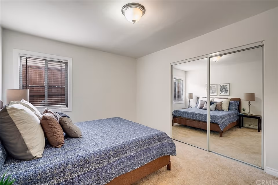 A bedroom with a bed covered in a blue patterned bedspread and multiple pillows. There is a window with blinds on the left wall and a mirrored sliding closet door on the right wall reflecting the bed and a nightstand with a lamp and small plant. The walls are painted white and the floor is carpeted.
