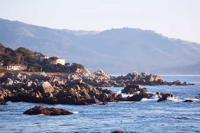 Rocky coastal shoreline with a few houses on the left and mountains in the background under a clear sky.