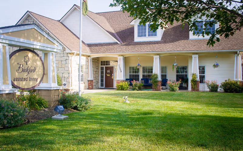 Exterior view of Bickford assisted living facility in Omaha Blondo, showing a well-maintained lawn, a sign with the facility name, and a building with a porch and seating area under a clear sky.