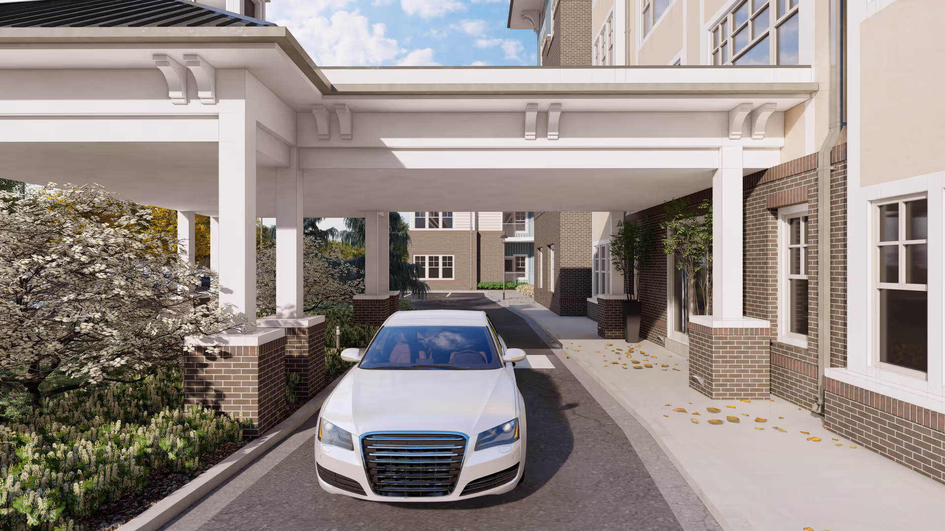 A white car parked under a covered driveway at the entrance of a brick and beige building with multiple windows. There are trees and shrubs on the left side and a sidewalk with some fallen leaves on the right side.