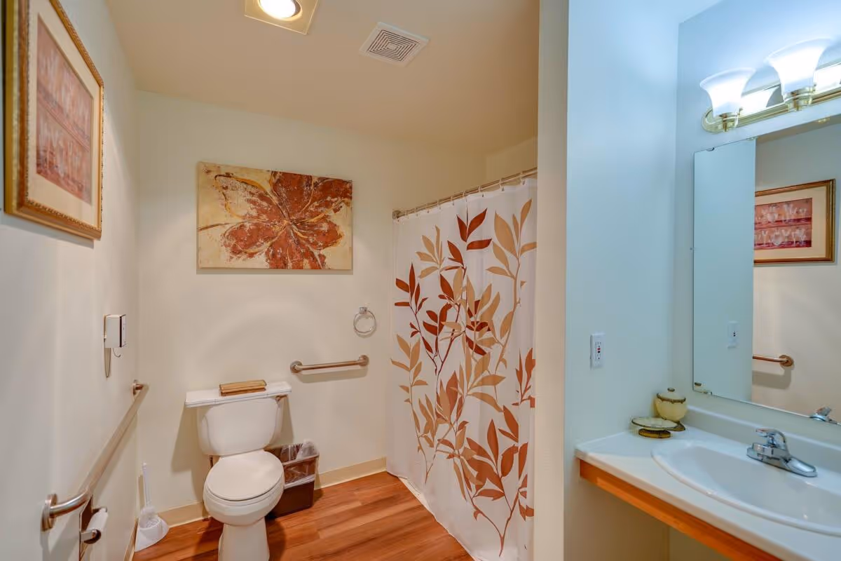 A clean and well-lit bathroom with a white toilet, a shower with a curtain featuring a leaf pattern in shades of brown and beige, a white sink with a mirror above it, and framed artwork on the walls. The floor is wooden, and there are safety grab bars installed near the toilet.