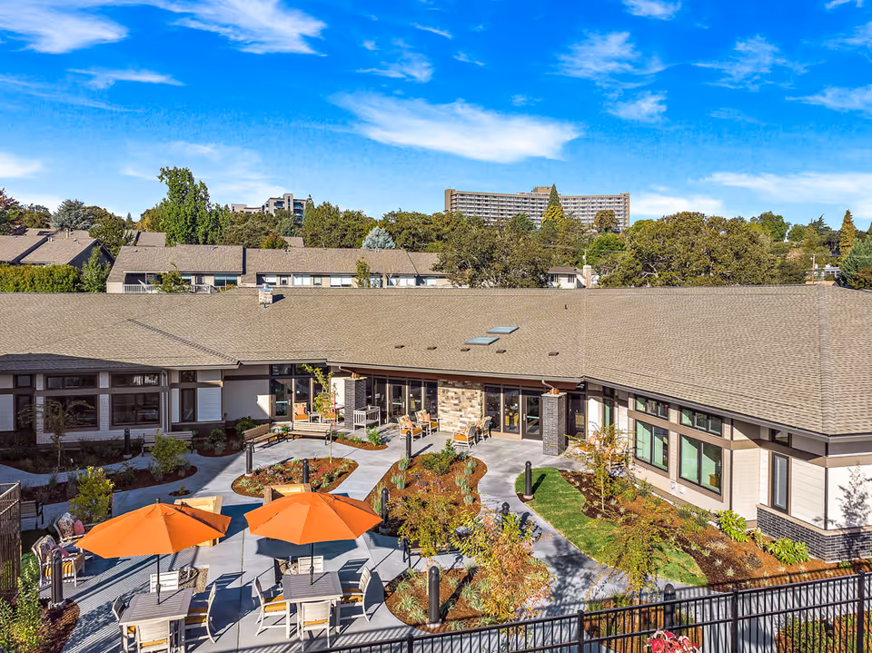 Aerial view of a landscaped courtyard at a senior living facility with seating areas, orange umbrellas, walkways, and surrounding single-story buildings under a blue sky.
