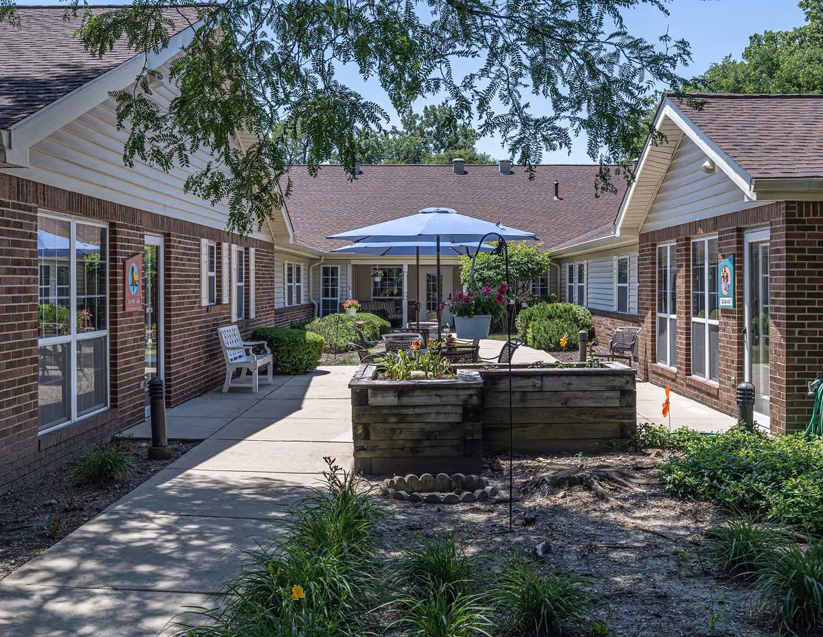 Outdoor courtyard area of Arden Courts - ProMedica Memory Care Community (Palos Heights) featuring a paved walkway, raised garden beds with flowers, patio tables with blue umbrellas, benches, and surrounding brick buildings with white trim under a clear blue sky.