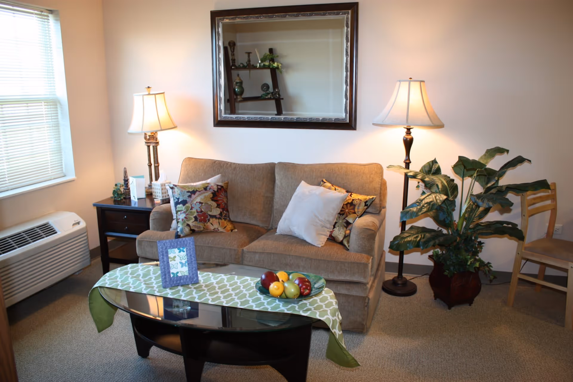 A cozy living room area with a beige couch adorned with three decorative pillows. There are two lamps on either side of the couch, one on a side table and the other a floor lamp. A large mirror hangs on the wall above the couch. A glass coffee table with a green patterned runner and a bowl of artificial fruit sits in front of the couch. A large potted plant and a wooden chair are positioned to the right. A window with blinds and an air conditioning unit are on the left side of the room.