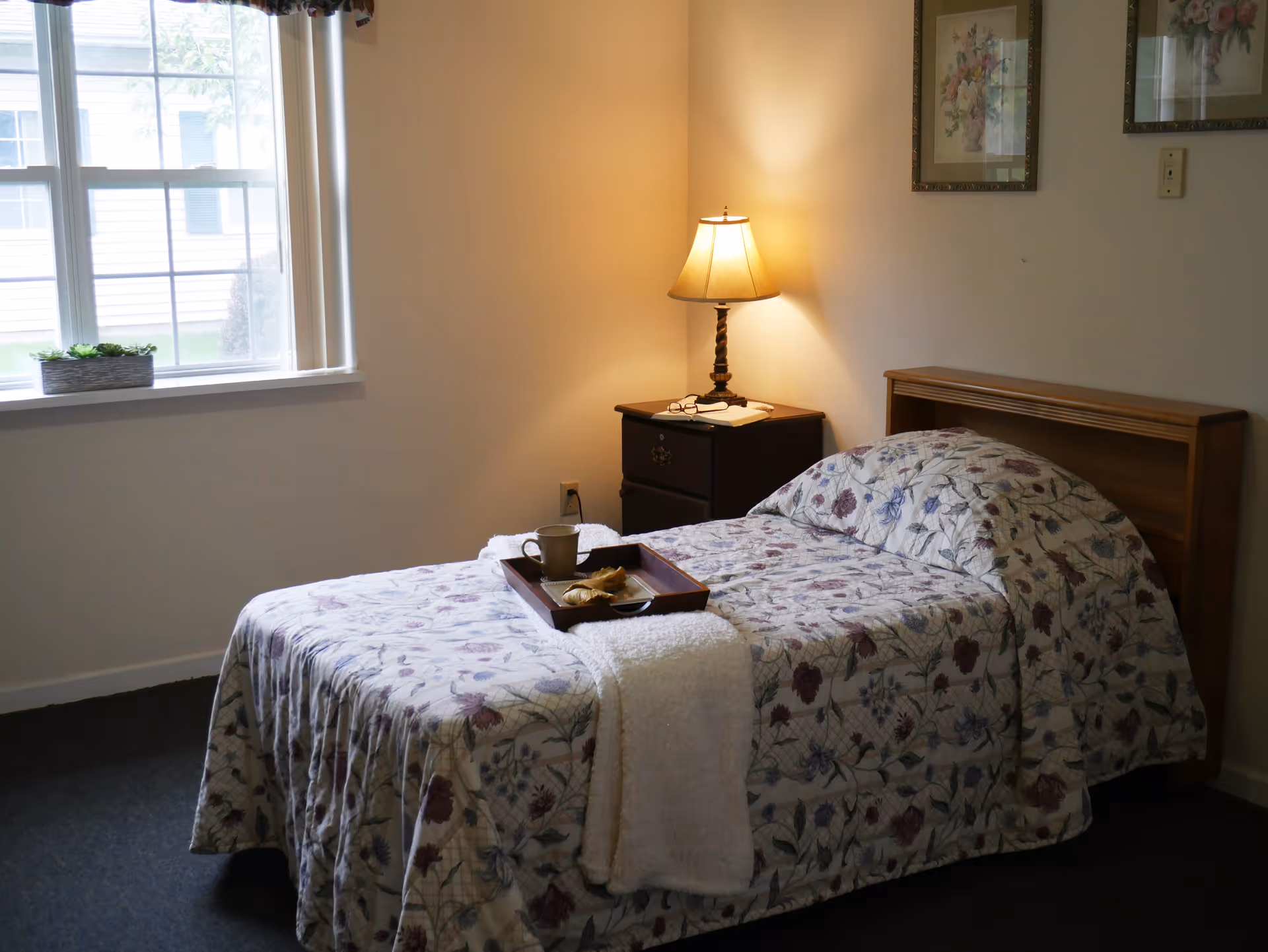 A small bedroom with a single bed covered in a floral bedspread, a nightstand with a lamp, a tray with a cup, and a window letting in daylight.