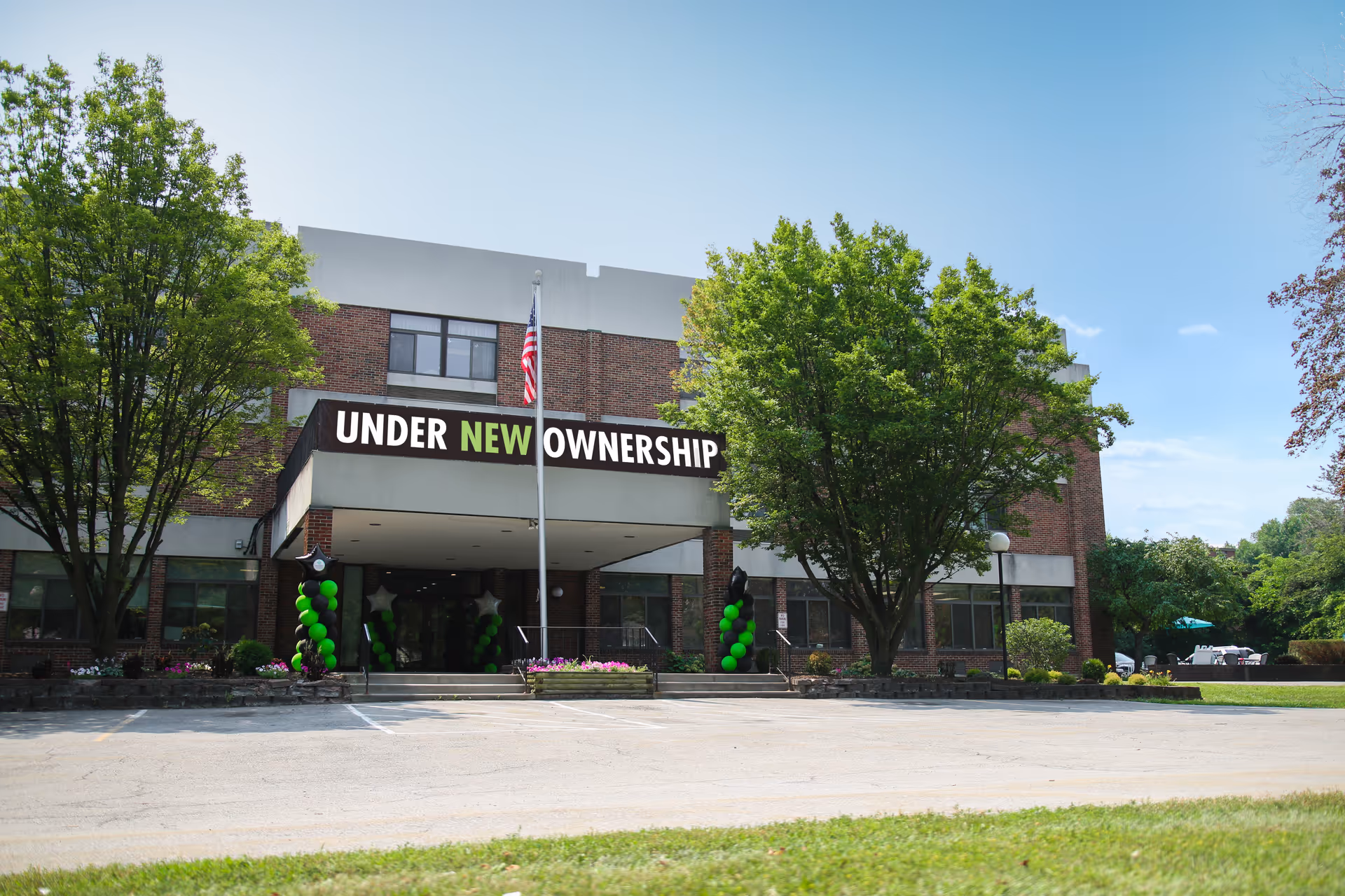 Brick senior living facility entrance with a banner reading 'UNDER NEW OWNERSHIP', an American flag, and green balloon decorations.