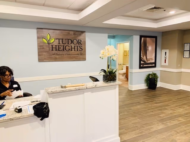 Reception area of Tudor Heights senior living facility with a white front desk, a woman sitting behind the desk, a wooden sign on the wall that reads 'Tudor Heights A Senior Community', a potted orchid on the desk, and a hallway leading to other rooms.