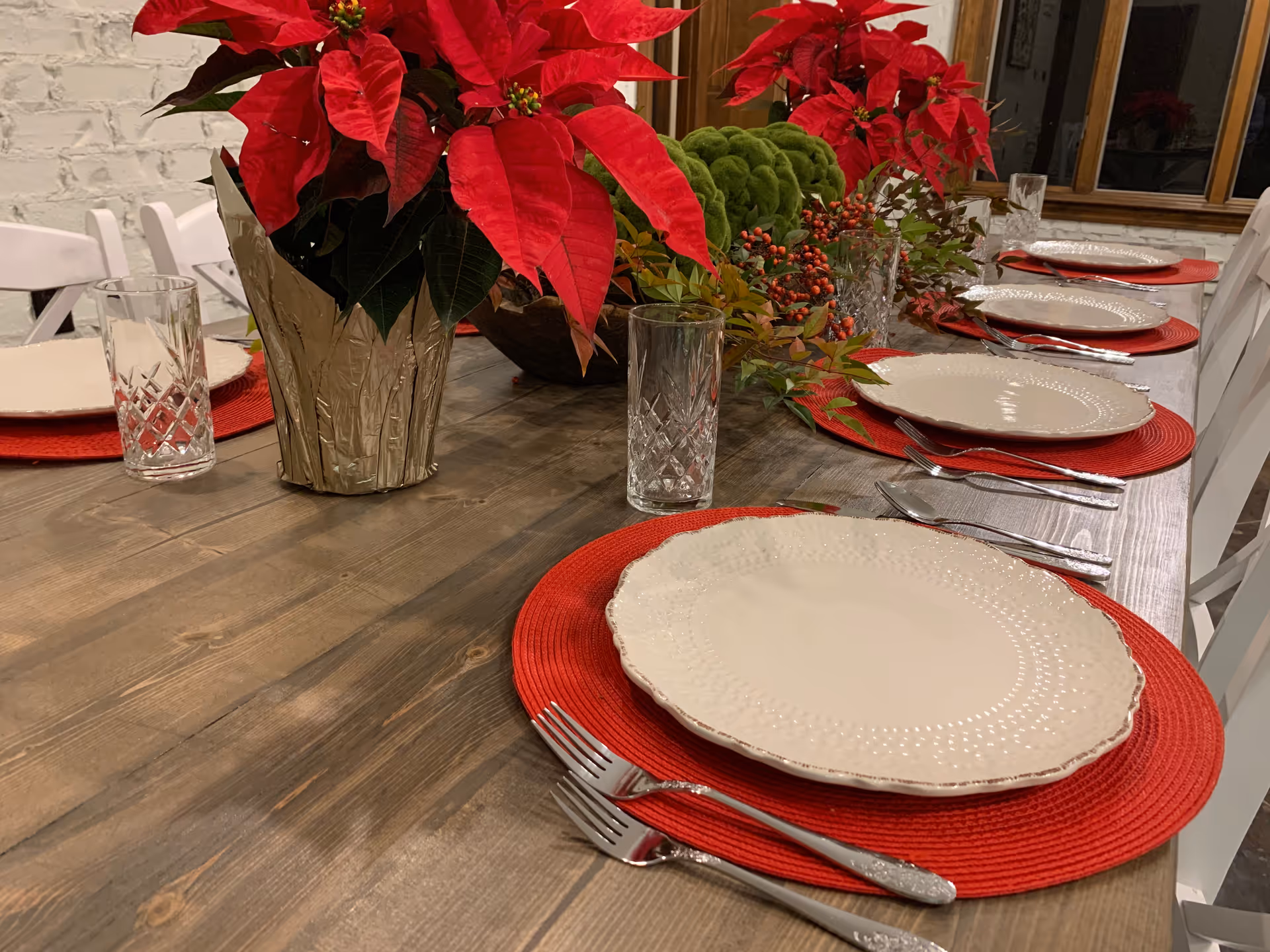 A wooden dining table set with white plates on red placemats, crystal glasses, cutlery, and poinsettia centerpieces.