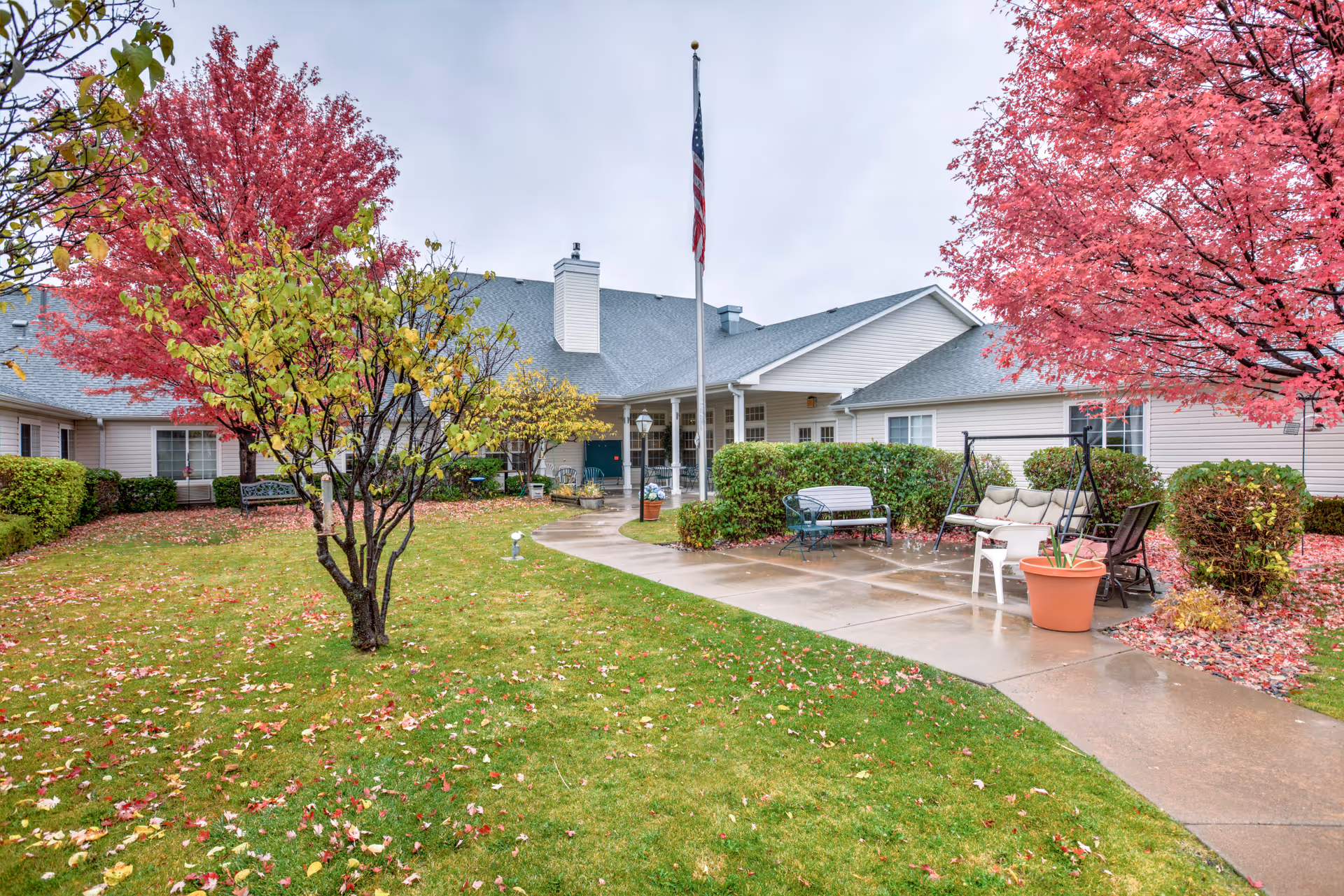 Courtyard of a one-story senior living building with a flagpole, benches and patio furniture surrounded by autumn trees and a wet walkway.