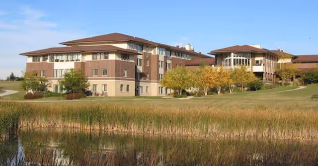 A large multi-story building with a brown and beige exterior surrounded by green grass, trees with autumn foliage, and a pond with tall reeds in the foreground under a clear blue sky.