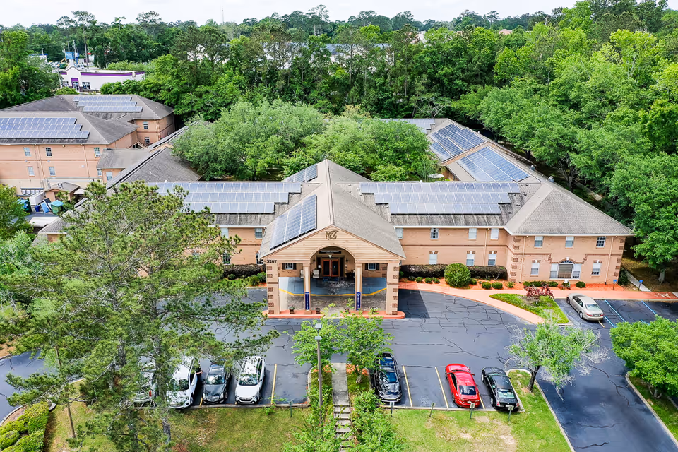 Aerial view of a brick senior living building with solar panels on the roof, a covered main entrance and a parking lot surrounded by trees.