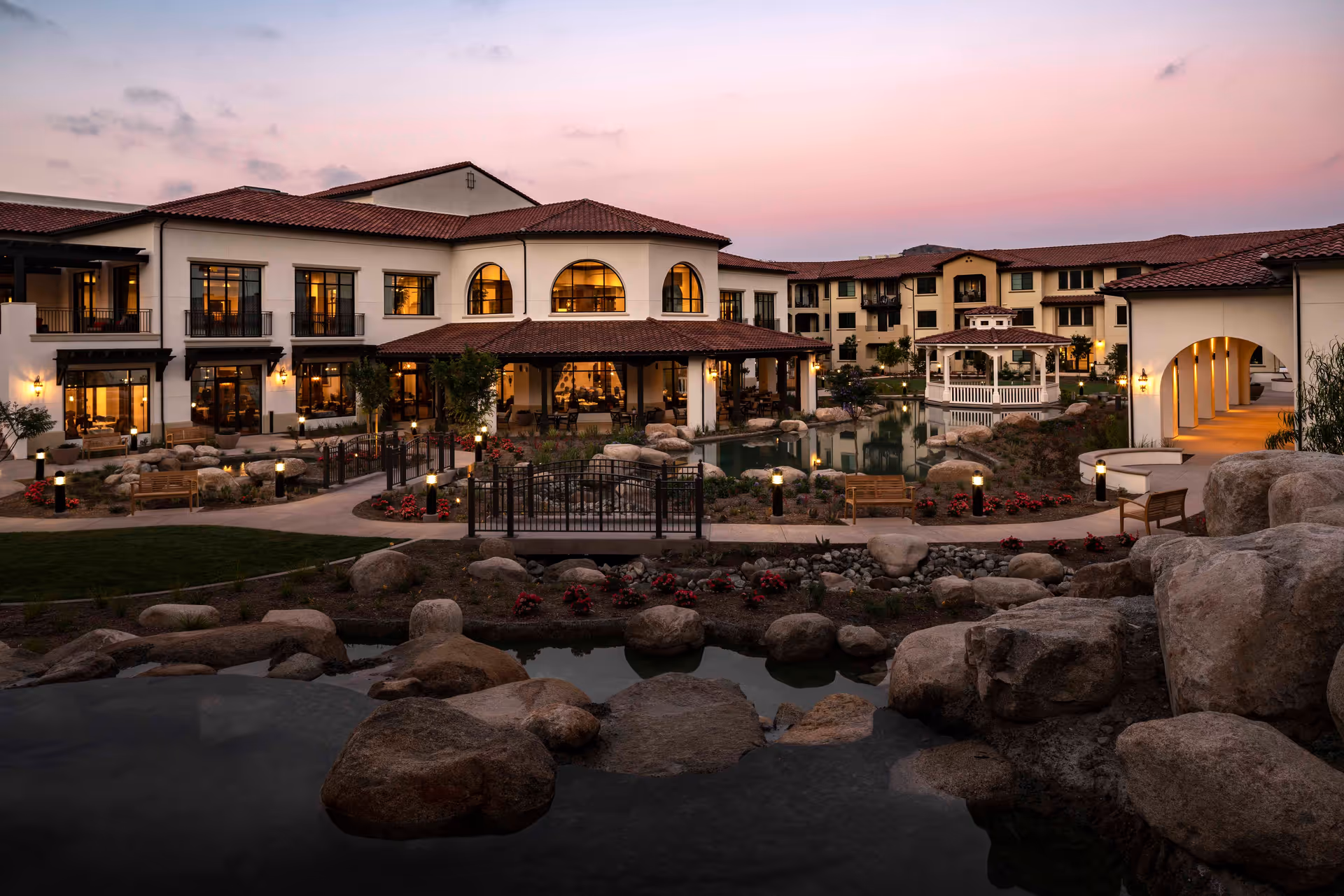 View of Reata Glen's illuminated courtyard and multi-story building at dusk, featuring a pond, gazebo, walkways, and landscaping.
