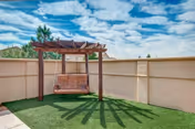 Outdoor area with artificial grass and a wooden pergola swing casting shadows on the ground, surrounded by a beige privacy wall under a partly cloudy blue sky.