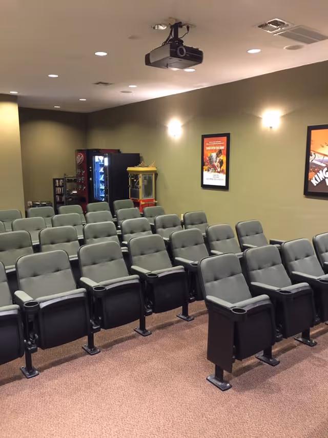 Small screening room with rows of theater seats, a ceiling projector, popcorn machine and vending machine.