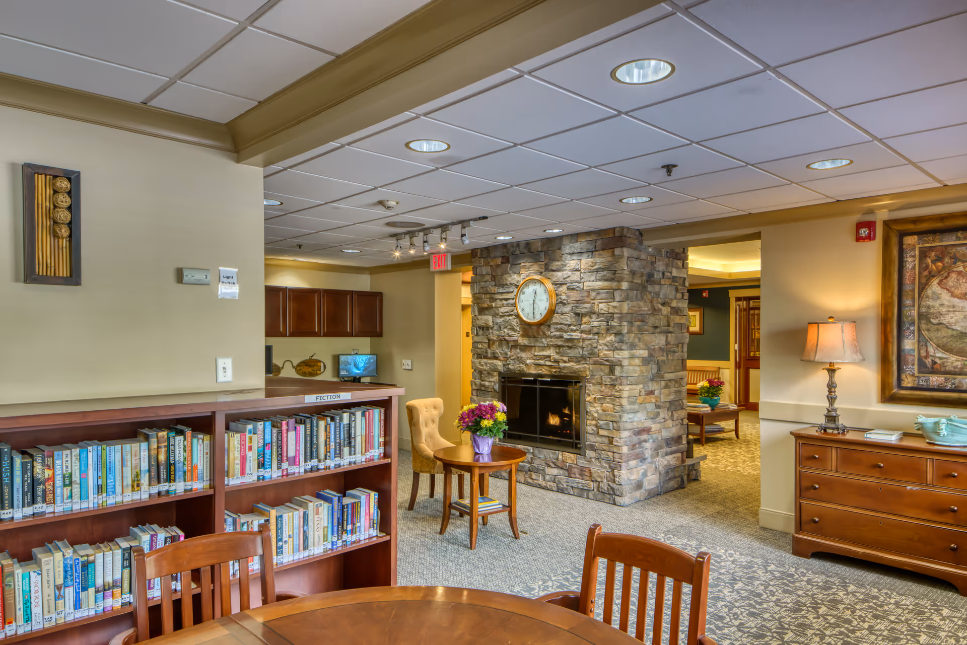 Interior of a senior living facility featuring a cozy common area with a stone fireplace and a clock above it. There is a wooden bookshelf filled with books labeled 'Fiction', a round wooden table with chairs, a small round table with a flower pot, and a cushioned chair. The room has a carpeted floor, beige walls, and a ceiling with recessed lighting. A wooden dresser with a lamp and decorative items is visible on the right side.
