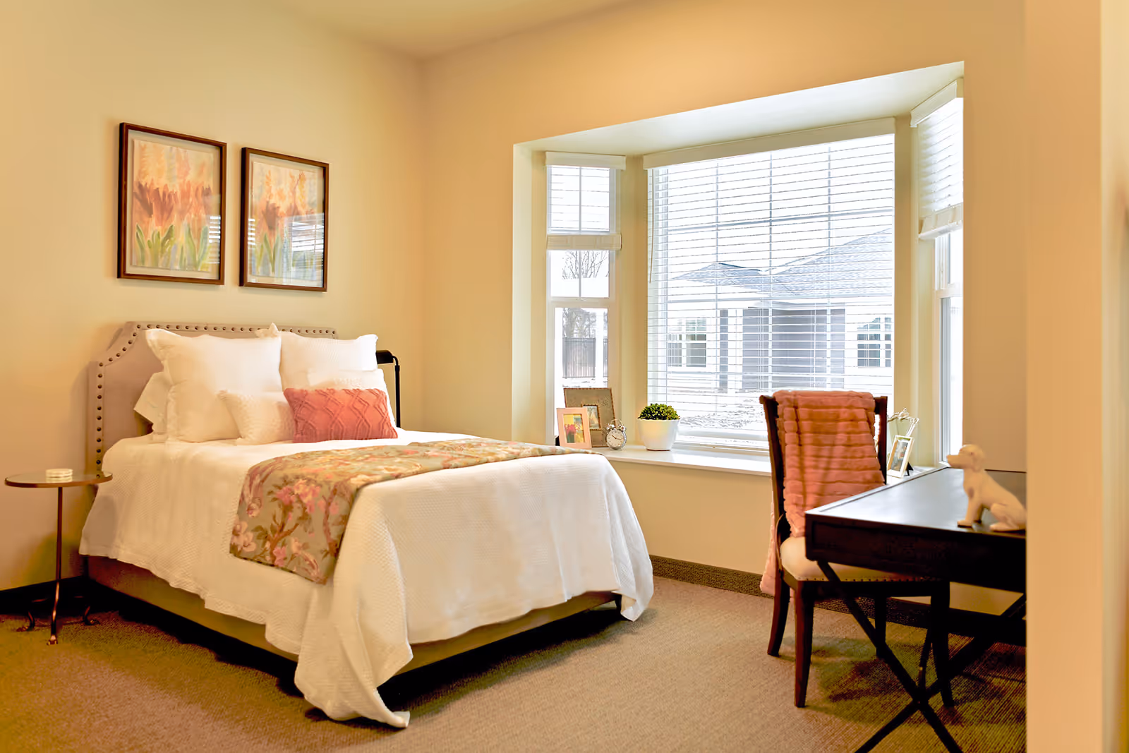 Cozy bedroom with a neatly made bed, decorative pillows, a bay window with blinds, and a small desk and chair.