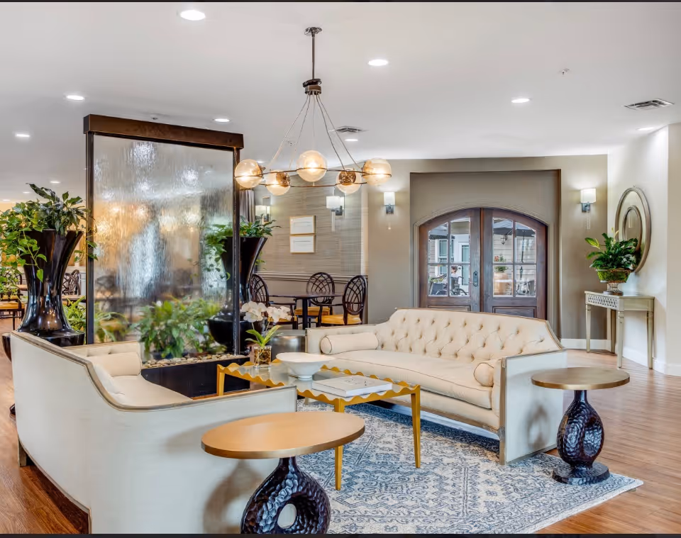 A bright and elegant living room area in a senior living facility featuring two cream-colored tufted sofas arranged around a gold coffee table on a patterned blue and white rug. There are two round side tables with dark sculpted bases, a large indoor water feature with plants, and a modern chandelier with globe lights hanging from the ceiling. In the background, there are dining tables and chairs, wall sconces, a console table with a plant and a round mirror, and double glass doors leading to another room.