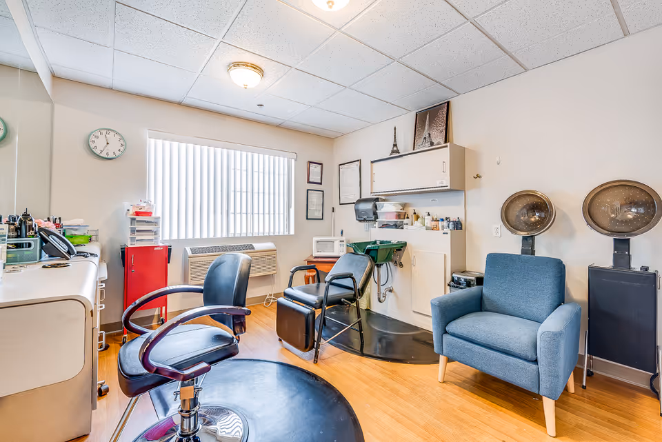 Small hair salon room with styling chairs, hooded hair dryers, a shampoo sink, and a blue armchair.