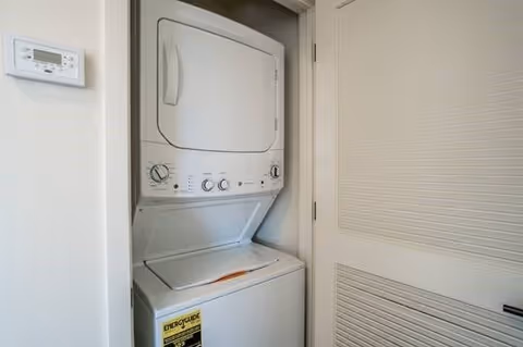 A compact stacked washer and dryer unit installed in a small closet space with a louvered door partially open. A thermostat is mounted on the adjacent wall.