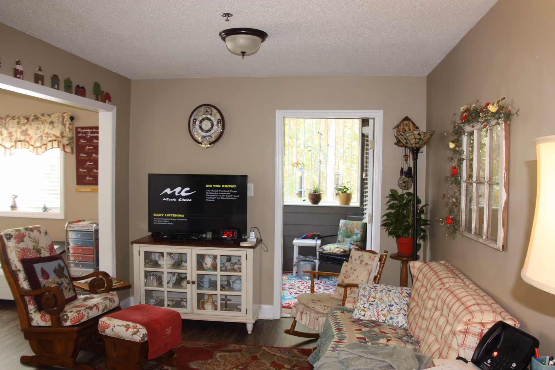 A cozy living room with a floral-patterned rocking chair and matching footstool, a plaid sofa with patterned pillows and a quilt, a wooden cabinet with glass doors displaying teapots and cups, a wall clock above the TV, and a doorway leading to a sunroom with potted plants and a chair. The walls are beige and decorated with a rustic window frame adorned with greenery and red birds, and a tall floor lamp stands in the corner.