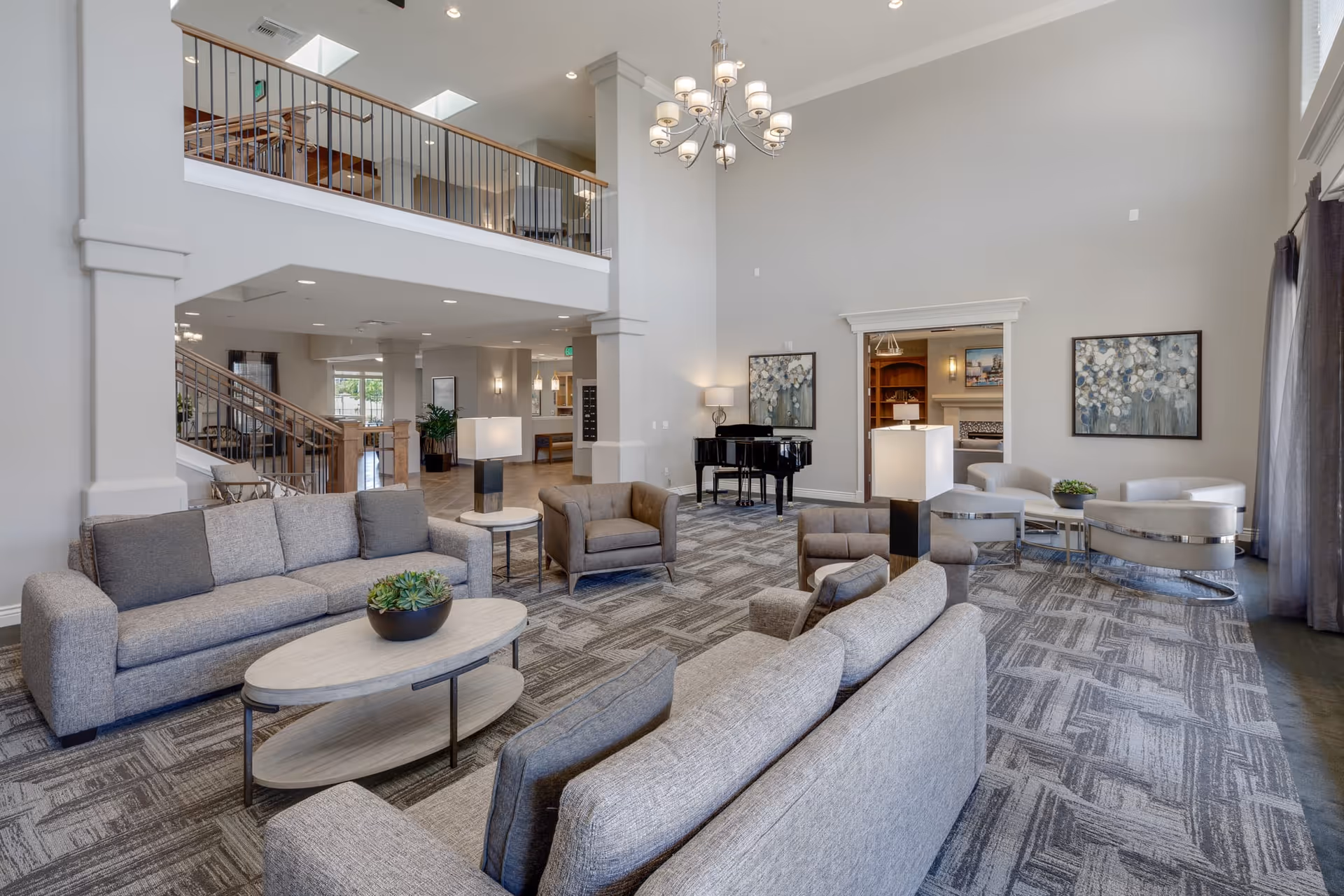 Spacious and modern senior living facility common area with multiple gray sofas and armchairs arranged around light wood coffee tables. The room features a high ceiling with a chandelier, large windows with curtains, a black grand piano, and decorative wall art. There is a staircase and an upper balcony visible in the background.