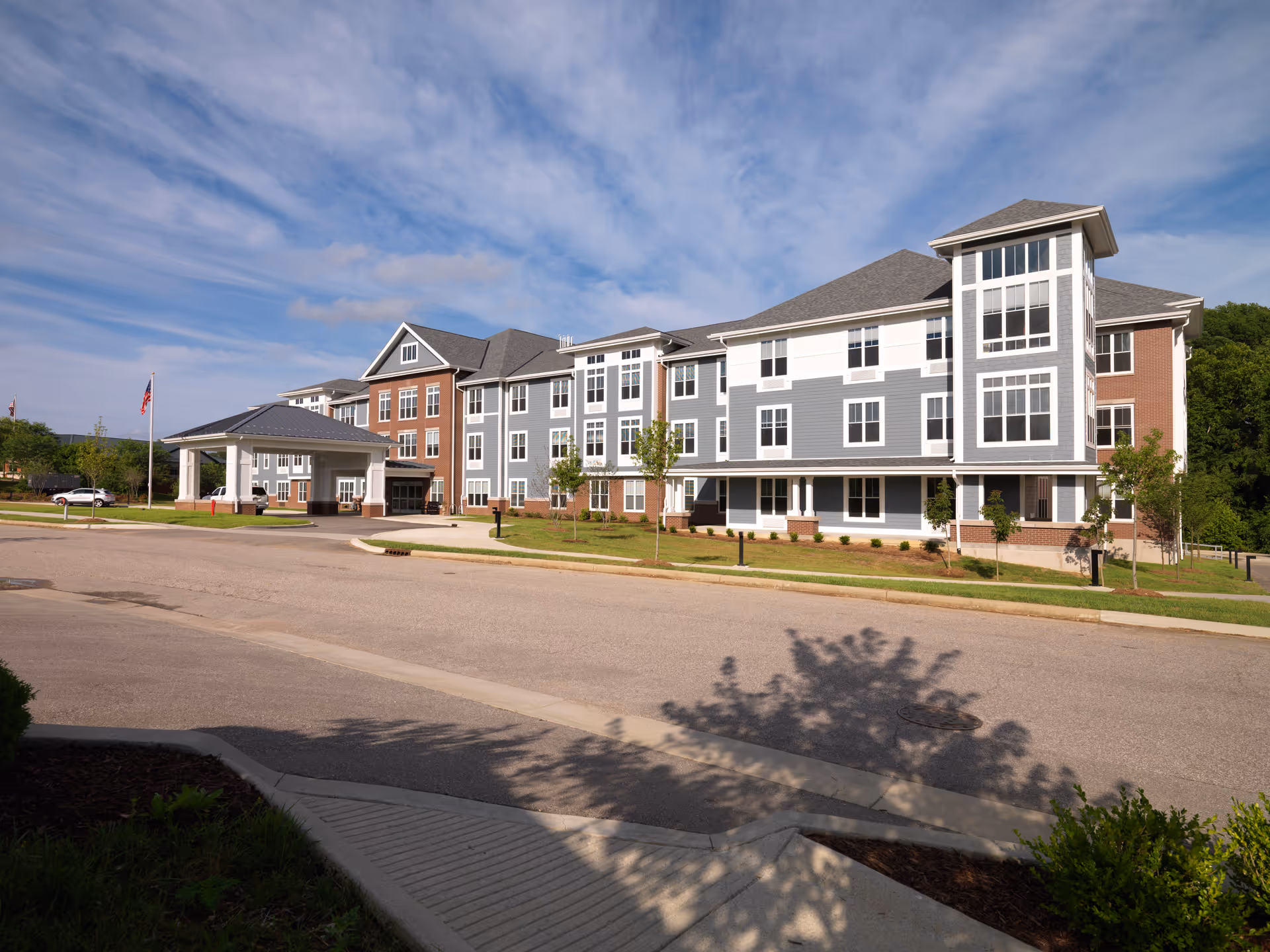 Exterior view of a large, modern senior living facility building with a covered entrance, multiple windows, and a mix of brick and gray siding under a partly cloudy sky.