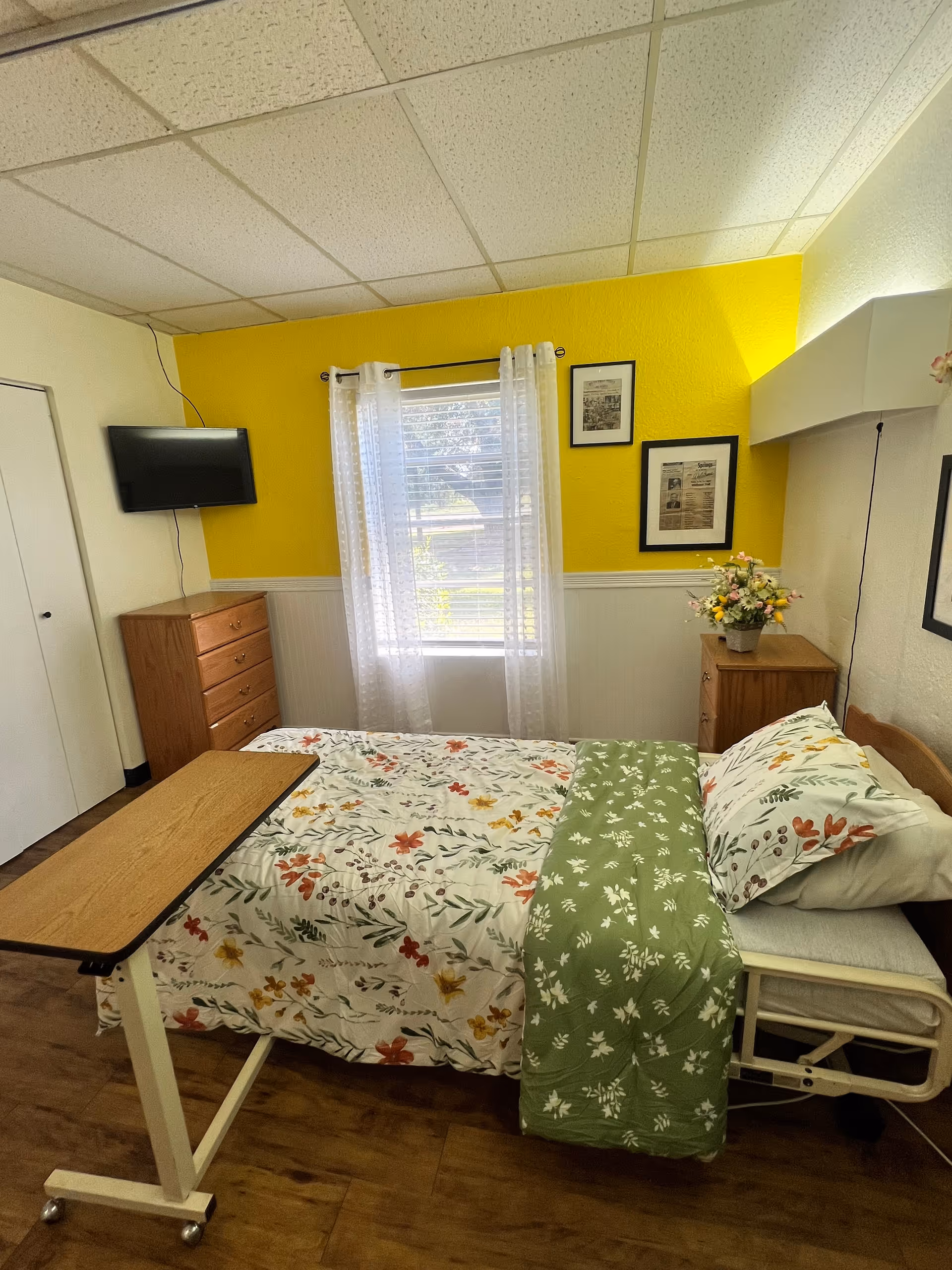 A small bedroom with a single bed covered in floral bedding and a green blanket. There is a wooden overbed table on wheels positioned over the bed. The room has a yellow accent wall with two framed pictures and a window with sheer white curtains. A wooden dresser and a small wooden nightstand with a flower arrangement are also visible. A flat-screen TV is mounted on the wall above the dresser.