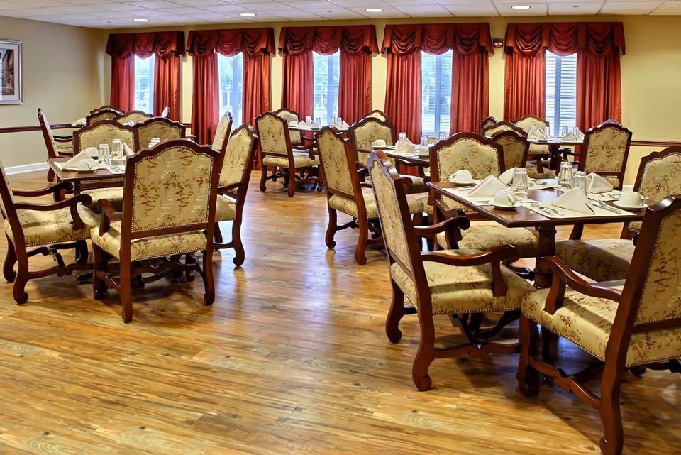 A dining room with multiple wooden tables and upholstered chairs arranged neatly. Each table is set with white napkins, cups, and glasses. The room has wooden flooring, yellow walls, and large windows with red curtains allowing natural light to enter.