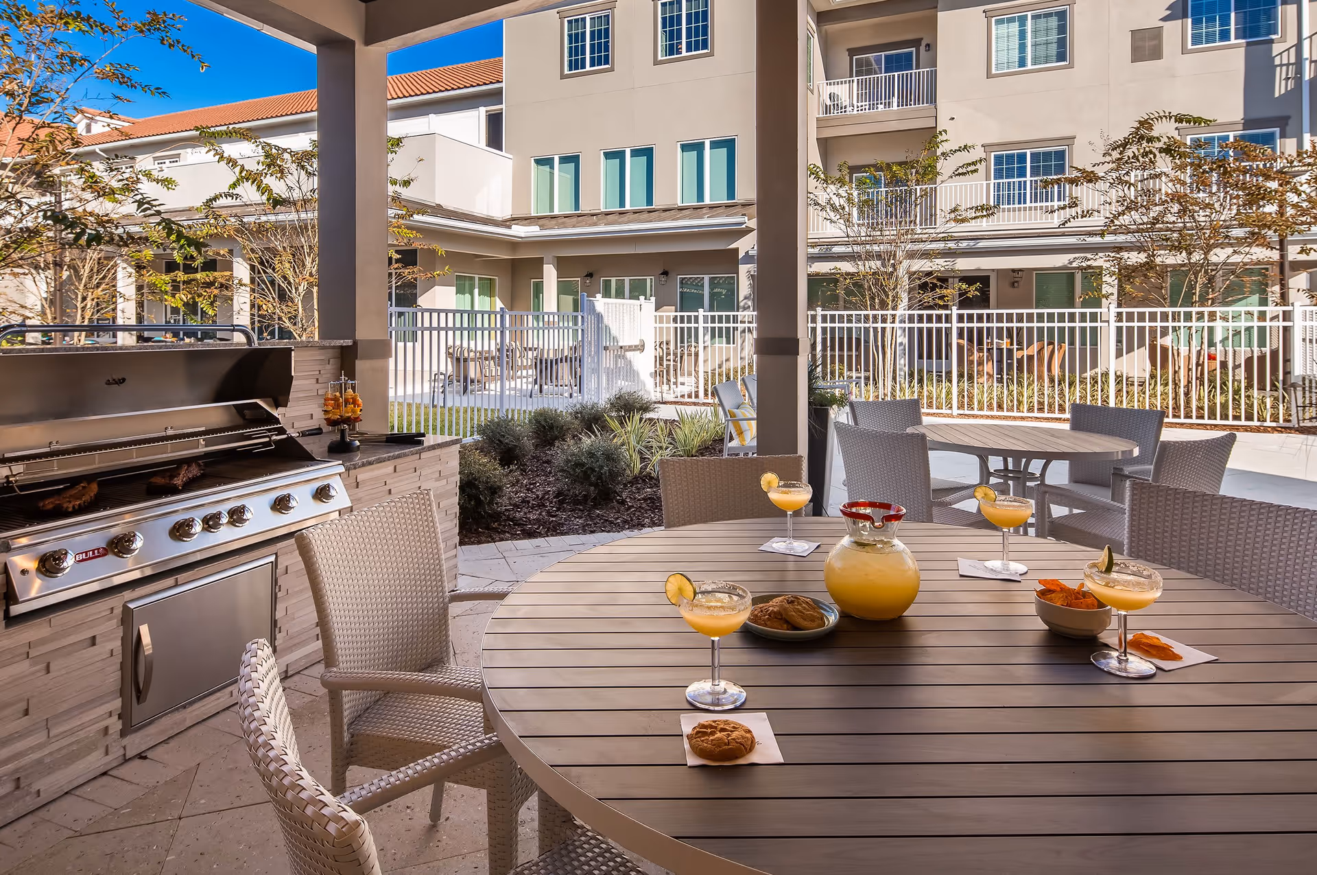Covered outdoor patio in a senior living courtyard with a built-in grill, round table set with drinks and snacks, and the apartment building in the background.
