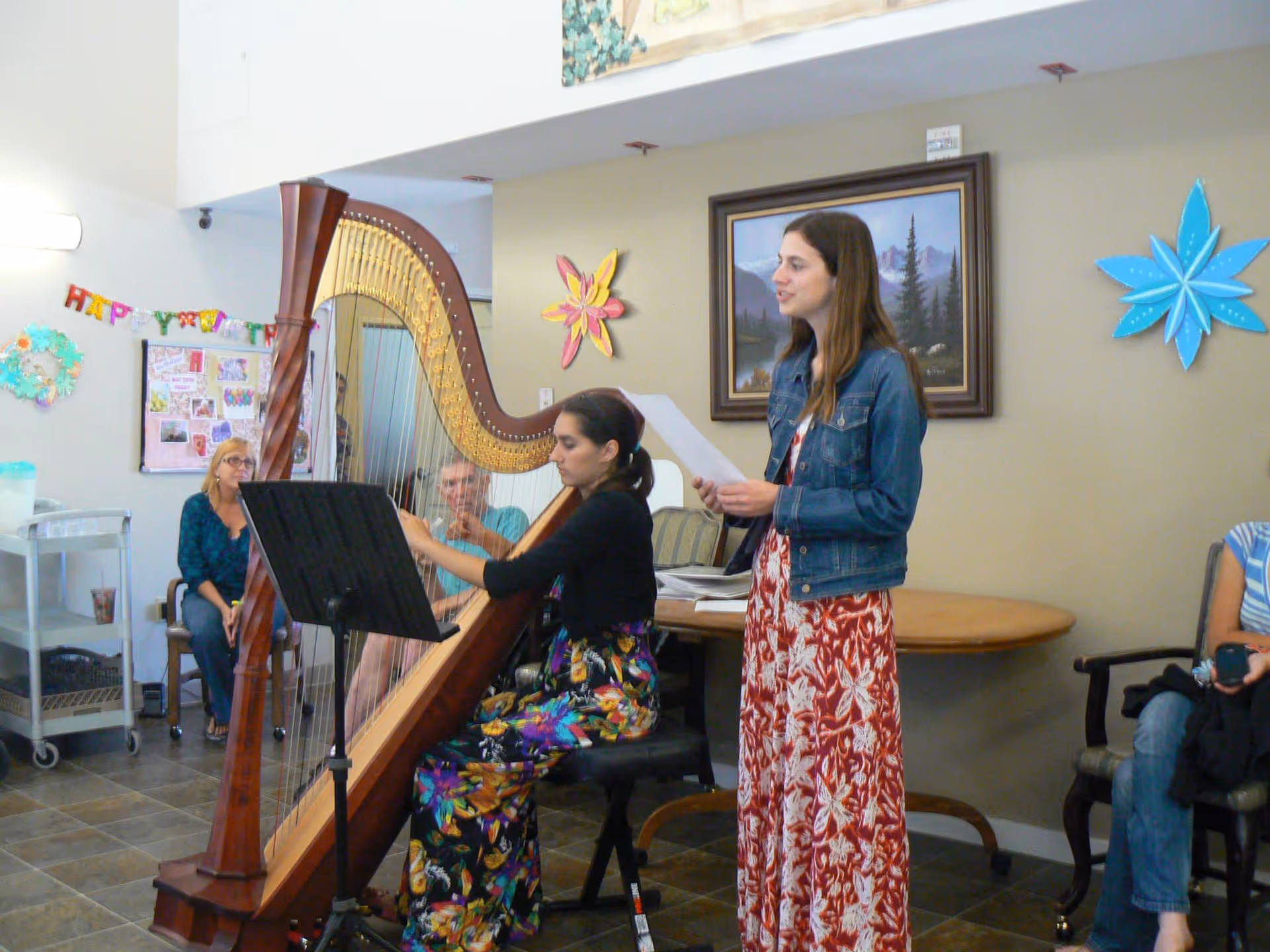 A woman seated and playing a large wooden harp while another woman stands beside her holding a sheet of paper, possibly singing. In the background, a few people are seated and watching. The room has a tiled floor, beige walls decorated with colorful paper flowers, and a framed landscape painting.
