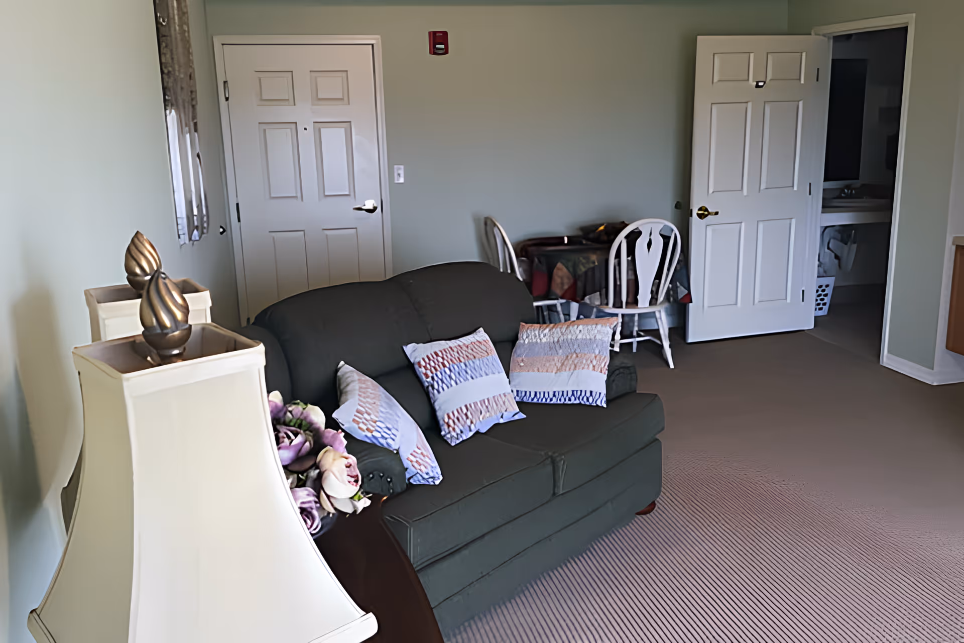 A cozy living room area with a dark green sofa adorned with three patterned pillows. To the left of the sofa is a white lamp with a decorative finial and a bouquet of purple flowers. In the background, there is a small table covered with a colorful tablecloth and two white wooden chairs. Two white doors are visible, one closed and one open, revealing a bathroom sink and laundry basket. The room has light green walls and a beige carpeted floor.