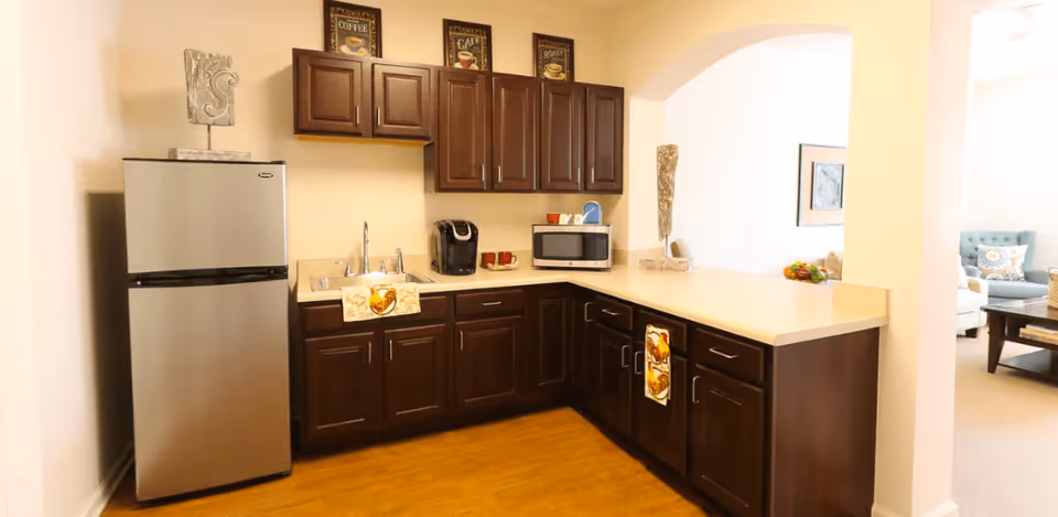 Small kitchen area with dark wood cabinets, a stainless steel refrigerator, sink, microwave, and a long countertop opening to a living room.