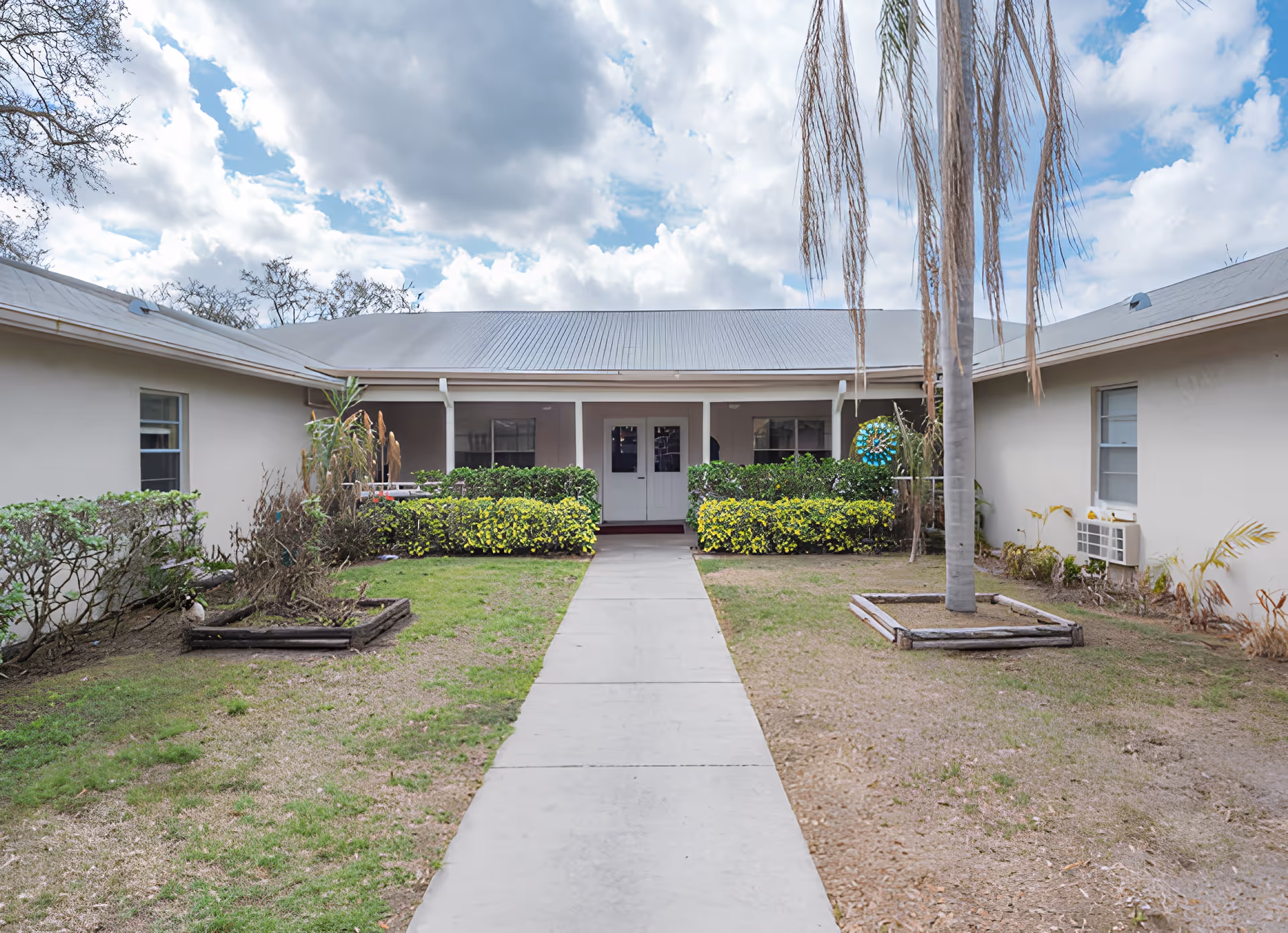 Exterior view of a single-story building with a gray metal roof and beige walls, featuring a central walkway leading to a double door entrance. The building is surrounded by trimmed bushes, a palm tree, and patches of grass under a partly cloudy sky.