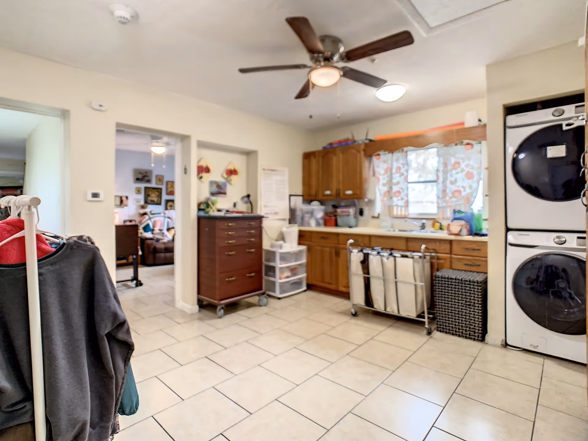 Interior view of a laundry room with tiled floor, wooden cabinets, a window with floral curtains, a stacked washer and dryer, a ceiling fan, and various laundry baskets and storage units. There is a clothing rack with clothes on the left side and an open doorway leading to another room with a person sitting on a couch.