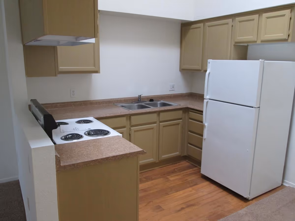 A small kitchen with beige cabinets, a white refrigerator, a white electric stove with four burners, a double stainless steel sink, and brown countertops. The floor is wood laminate, and the walls are painted white.