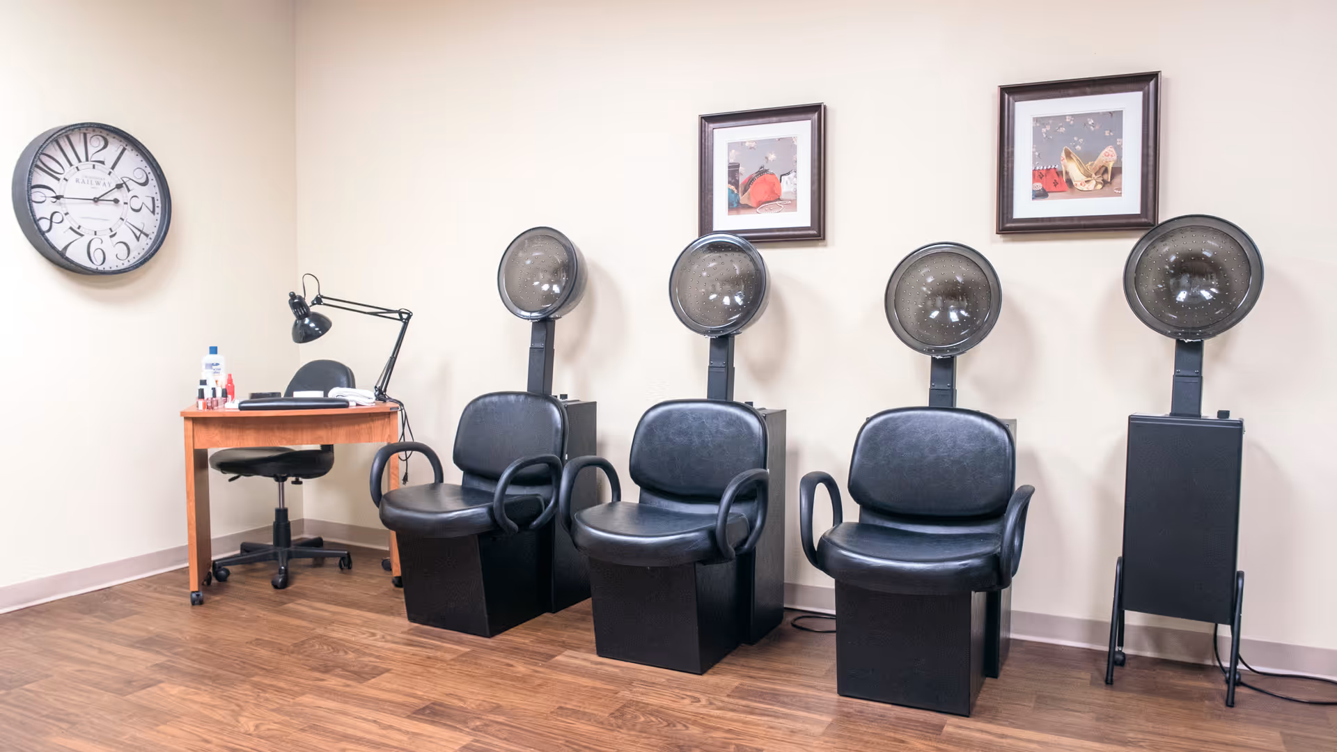 Interior salon area with four black hooded hair dryers and chairs against a beige wall, a small desk with a lamp and a large wall clock.