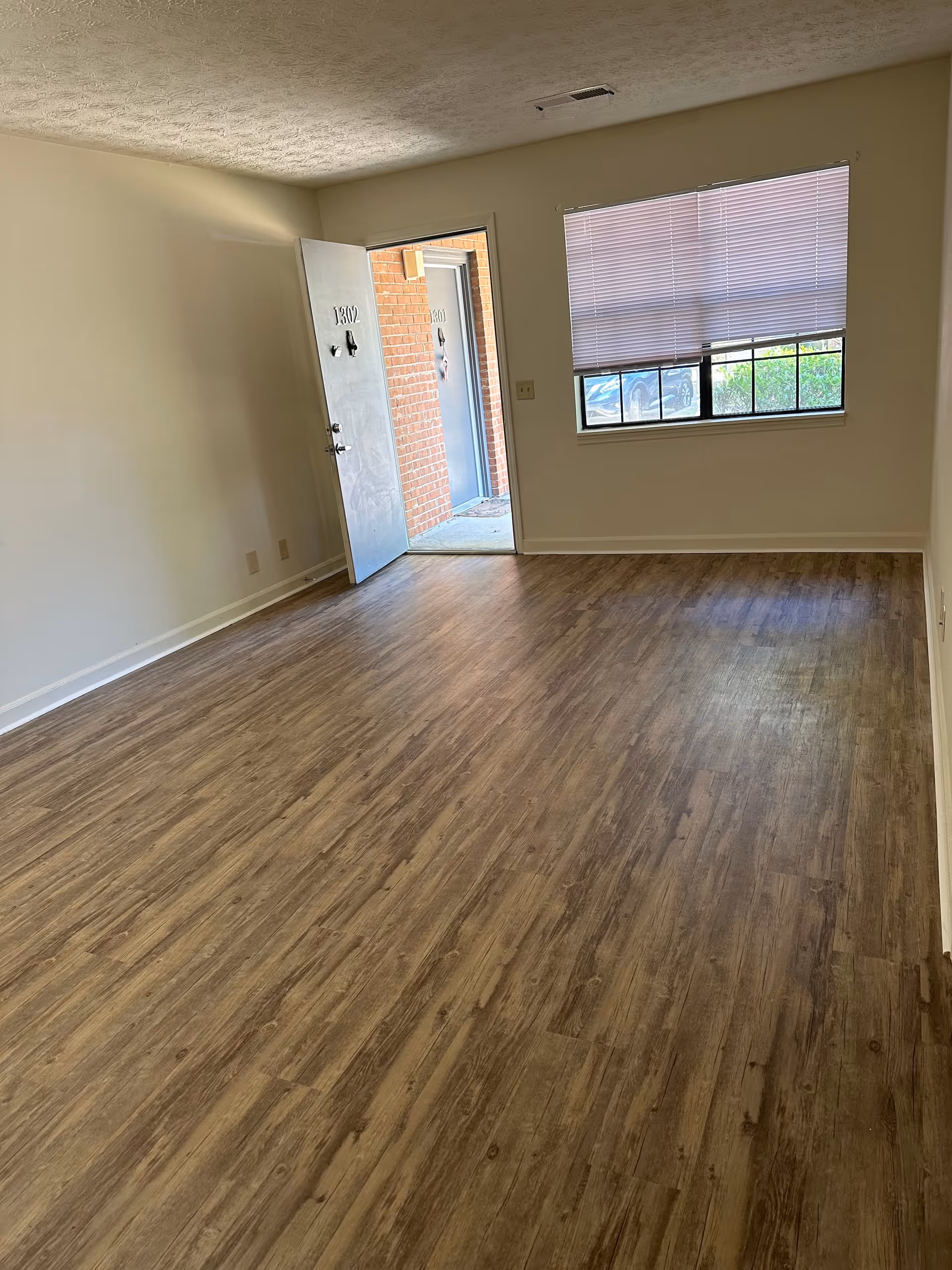 Empty room with wood-patterned flooring, a window with closed blinds, and an open door showing a brick exterior and another door outside.