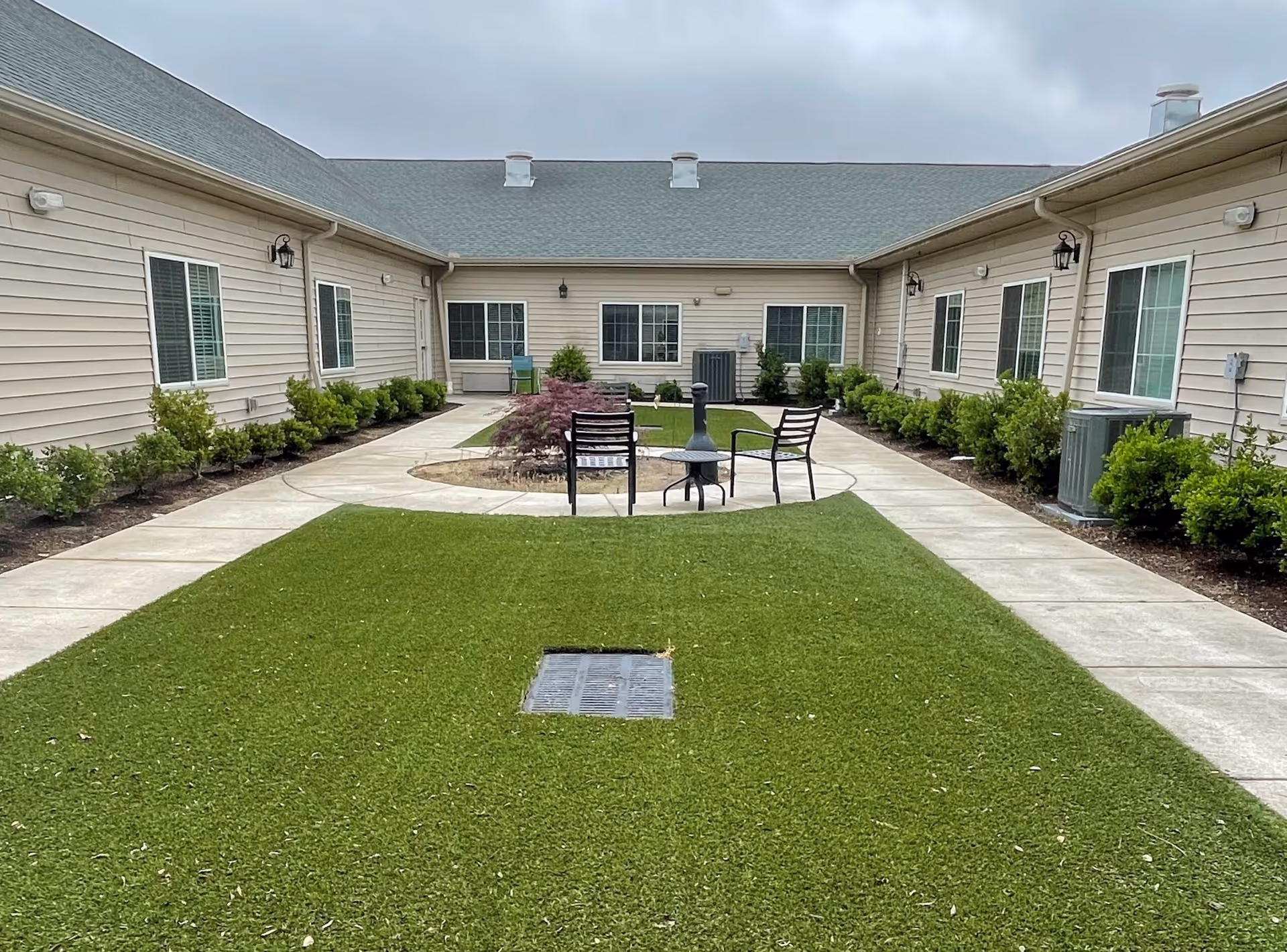 Outdoor courtyard area with green grass, a circular paved section with two black chairs and a small table, surrounded by beige buildings with multiple windows and small bushes along the walkways.
