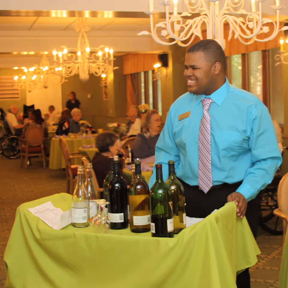 A smiling male staff member in a turquoise shirt and striped tie stands behind a table covered with a green tablecloth, which holds several bottles of wine and glasses. In the background, elderly residents are seated at tables in a warmly lit dining room with chandeliers and large windows.