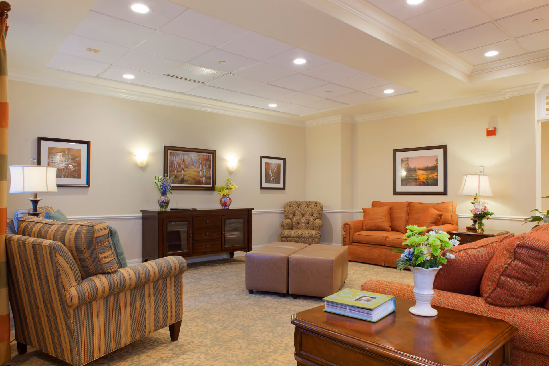 A cozy senior living facility lounge with multiple upholstered chairs and sofas in warm tones, a wooden coffee table with a vase of flowers and a book, framed artwork on the walls, and soft lighting from lamps and wall sconces.