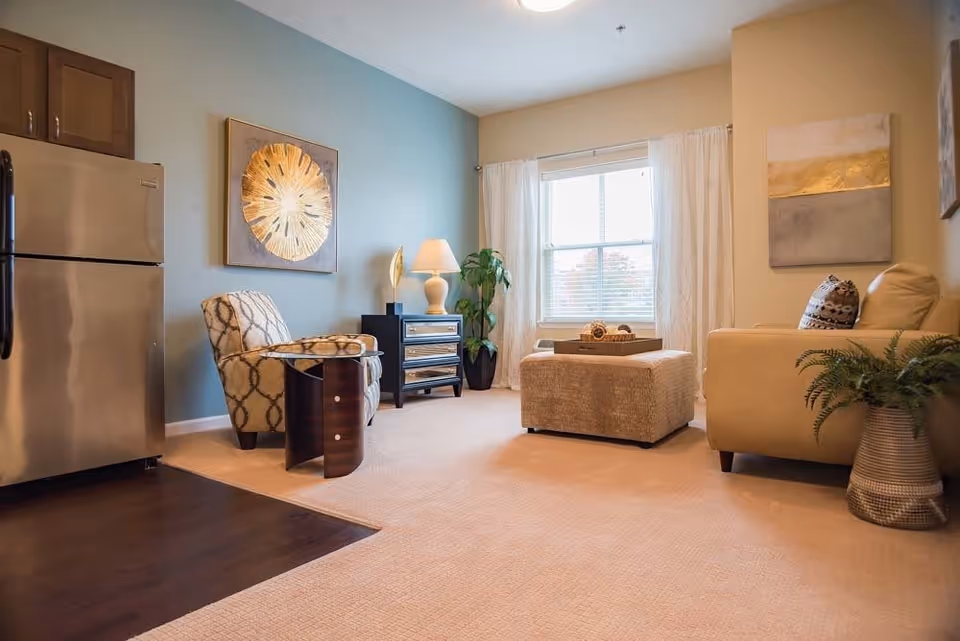 A cozy living room area with a beige carpet, a patterned armchair, a beige sofa with decorative pillows, a large ottoman, a side table with a lamp, a potted plant, and a window with white curtains letting in natural light. A stainless steel refrigerator is partially visible on the left side, indicating an adjoining kitchen area.