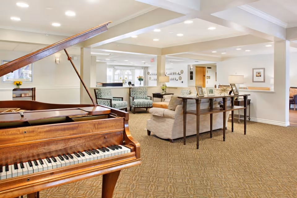 A spacious and well-lit living room area featuring a wooden grand piano in the foreground. The room has beige carpeting and neutral-colored walls with recessed lighting. There are several upholstered chairs and sofas arranged around wooden tables, some with framed photos and decorative items. The background shows a reception desk area and additional seating with large windows allowing natural light.