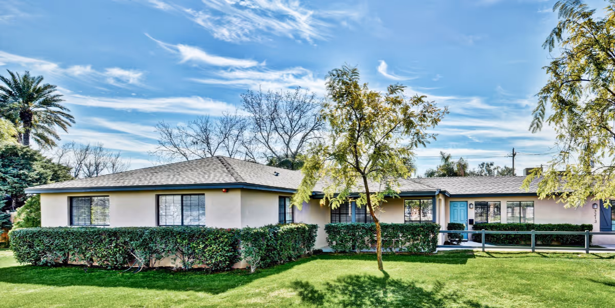Single-story assisted living building with trimmed hedges, a green lawn, trees and a blue front door under a bright sky.